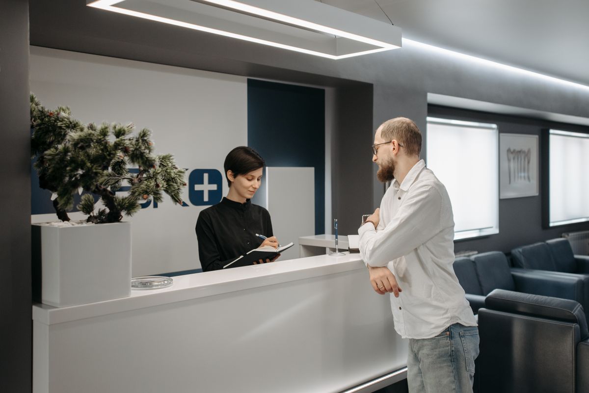 Receptionist at a small medical practice on the phone while a doctor reviews a patient file behind her